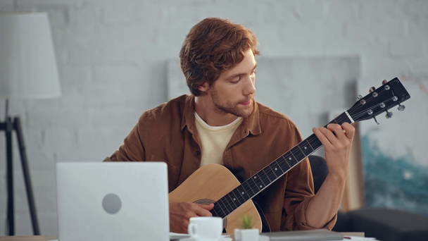 κοκκινομάλλα άνθρωπος παίζει ακουστική κιθάρα, ενώ κοιτάζοντας την οθόνη του φορητού υπολογιστή - Πλάνα, βίντεο