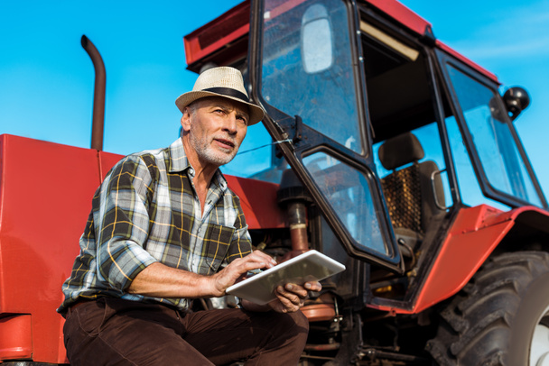 agricultor senior en sombrero de paja utilizando tableta digital cerca de tractor rojo
  - Foto, Imagen