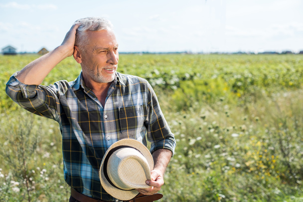 笑顔の先輩農家が髪に触れ、畑で麦わら帽子を持っている  - 写真・画像