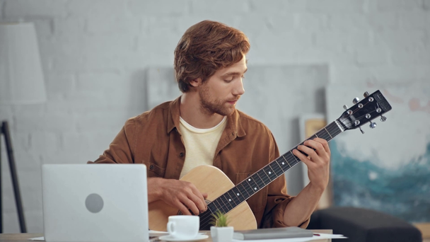 thoughtful redhead man playing acoustic guitar near laptop - Video