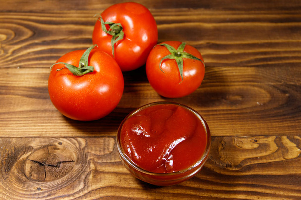Tigela de vidro de ketchup ou molho de tomate e tomates frescos maduros em mesa de madeira
 - Foto, Imagem
