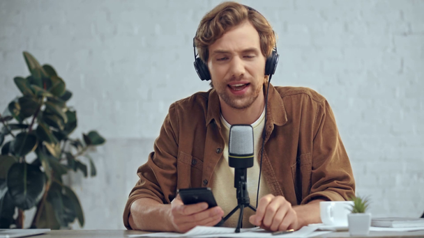 man in headphones with mic broadcasting and using smartphone - Felvétel, videó