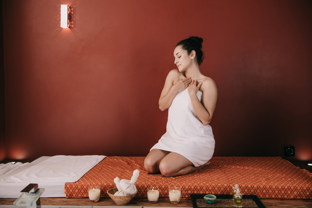 attraktive Frau mit geschlossenen Augen im Handtuch auf Massagematte sitzend  - Foto, Bild