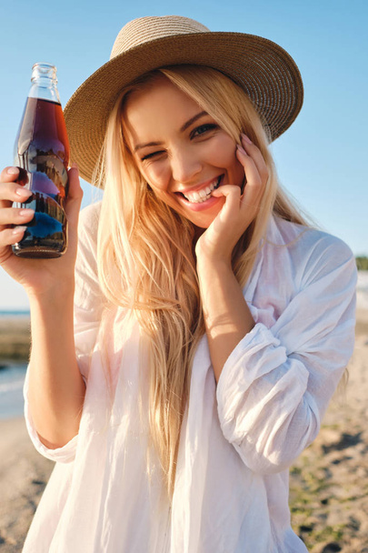 Young cheerful blond woman in white shirt and hat holding bottle of soda water joyfully looking in camera by the sea - Photo, image