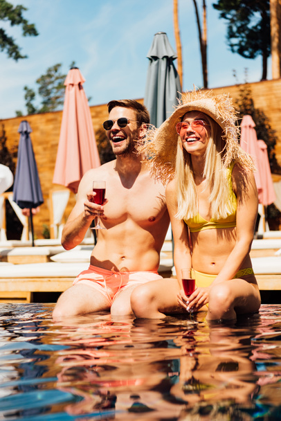 sexy couple holding glasses of red wine and smiling in swimming pool - Valokuva, kuva
