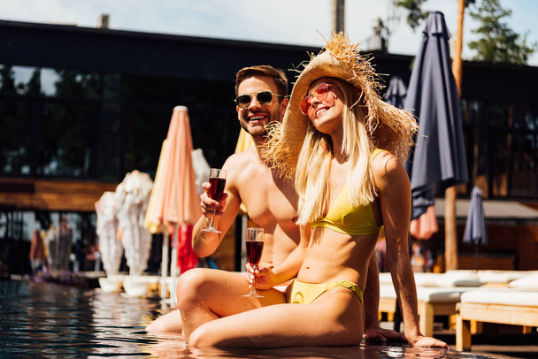 sexy couple holding glasses of red wine and smiling in swimming pool - Fotografie, Obrázek