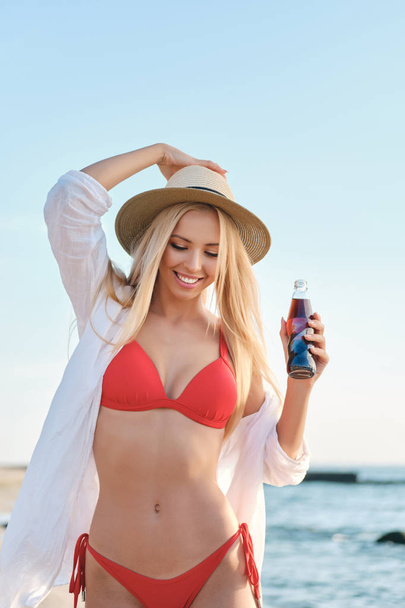 Молода красива усміхнена блондинка в купальнику і капелюсі тримає скляну пляшку з содовою водою мрійливо йде біля моря
 - Фото, зображення
