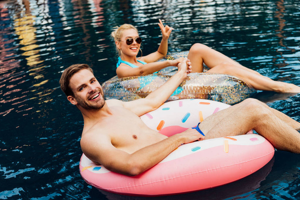 улыбающаяся пара, держащаяся за руки, лежа на плавательных кольцах в бассейне
 - Фото, изображение