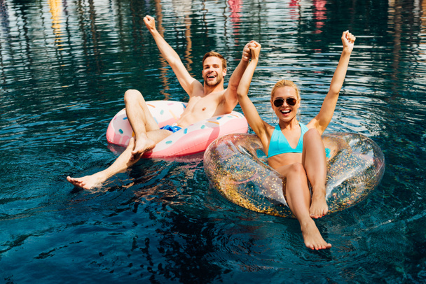 happy couple holding hands while lying on swim rings in swimming pool - Photo, Image