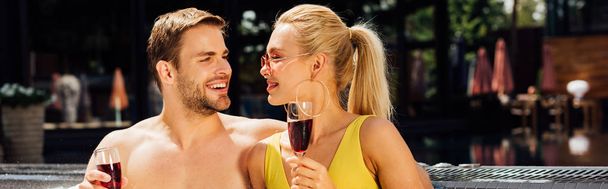 panoramic shot of sexy couple holding wine glasses with red wine and looking at each other at resort - Photo, Image