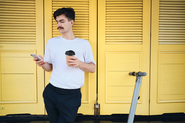 Jovem bigode homem de pé com café para ir e scooter pensativo usando celular na rua da cidade
  - Foto, Imagem