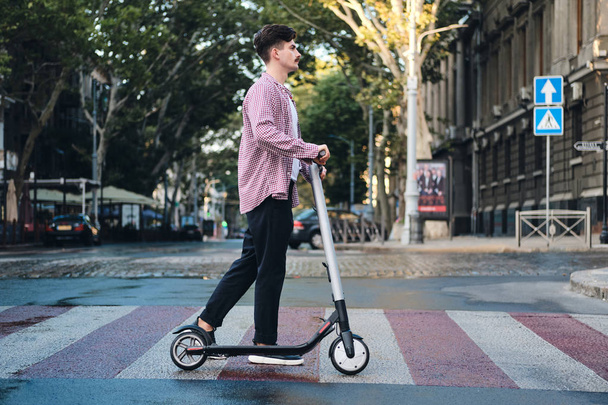 Side view of young man with e-scooter going on pedestrian crossing on city street  - Fotografie, Obrázek