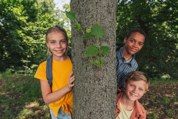 szczęśliwy wielokulturowy dzieciaki uśmiechający się w pobliżu drzewa  - Zdjęcie, obraz