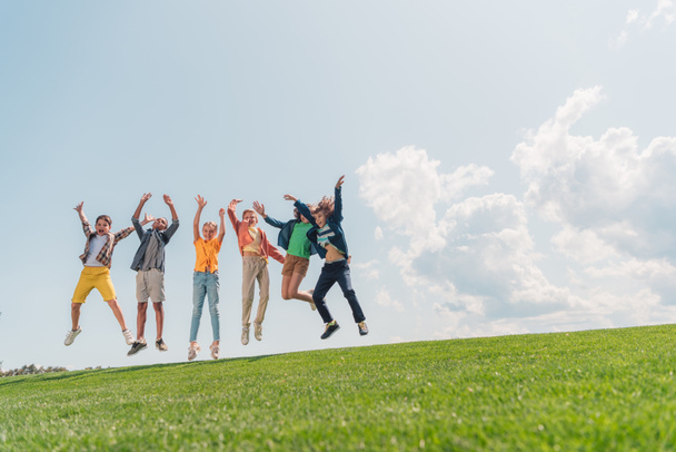 cheerful multicultural kids jumping and gesturing against blue sky  - Fotoğraf, Görsel
