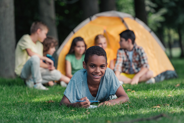 selective focus of happy african american boy holding pencil near notebook and friends in camp  - Valokuva, kuva
