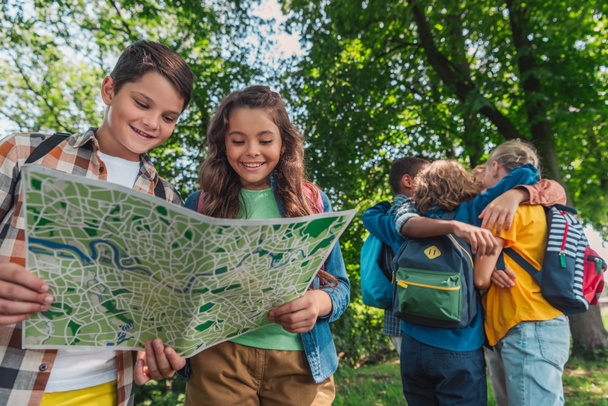 selective focus of happy kids looking at map near multicultural friends hugging in park  - Foto, Imagem