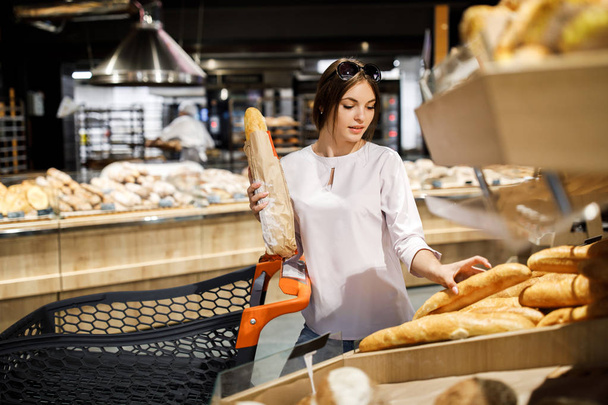 Young pretty girl is shopping in a big store. The girl buys groceries at the supermarket. The girl chooses a fragrant pastry - Fotoğraf, Görsel