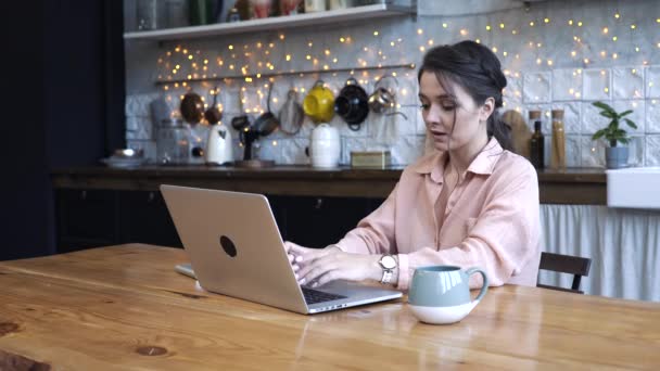 Concentrated young woman sitting in the kitchen at wooden table and typing on her personal silver laptop with the cup of tea or coffe. Stock footage. Modern decorated home interior on the background - Metraje, vídeo