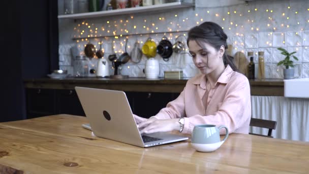 Concentrated young woman sitting in the kitchen at wooden table and typing on her personal silver laptop with the cup of tea or coffe. Stock footage. Modern decorated home interior on the background - Séquence, vidéo