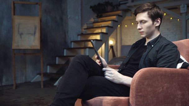 Zbliżenie młodego przystojnego mężczyzny w czarnej koszulce i dżinsy patrząc na cyfrowy tablet siedząc w dużym wygodnym fotelu w domu. Materiały stockowe. Nowoczesny wystrój wnętrz na tle - Zdjęcie, obraz