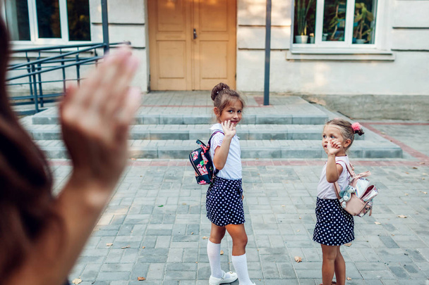 Молодая мать машет своим дочерям перед занятиями на открытом воздухе начальной школы провожает их. Семья прощается
. - Фото, изображение