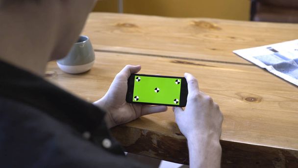 Blick von der Schulter eines jungen Mannes in schwarzem Hemd, der an einem Holztisch sitzt und zu Hause auf den grünen Bildschirm schaut. Archivmaterial. Smartphone neues Technologie-Konzept - Foto, Bild