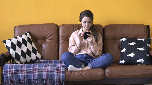 View of young pretty woman in pink shirt and jeans sitting on the brown couch with pillows and typing on her smartphone. Stock footage. Woman relaxing at home - Photo, image