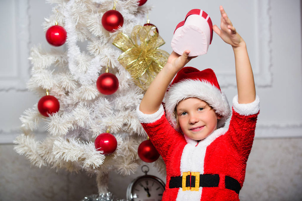 Christmas santa claus costume for child. Christmas party with santa costume. Christmas tree ideas for kids. Boy kid dressed as santa with red hat hold gift box near christmas tree - Fotó, kép