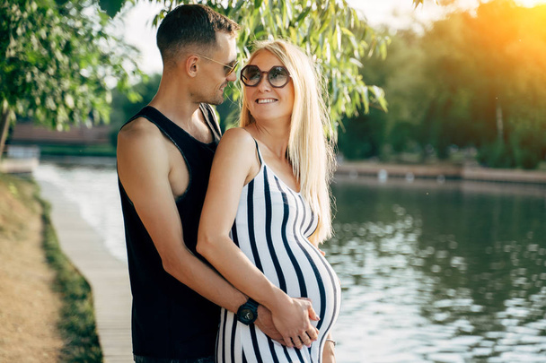 Photo of pregnant blonde woman and men standing on bank of river on summer day - Foto, imagen