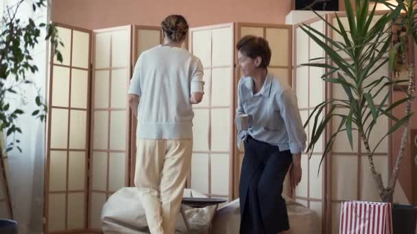 Two adult women with cups of coffee or tea sitting at the small table on comfortable pillow chairs. Two elegant mature girlfriends relaxing at home, talking and sharing news. Slow motion. - Materiał filmowy, wideo