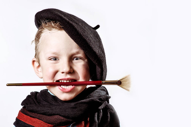boy artist in black beret, scarf and with a brush in his mouth on a light background - Foto, imagen