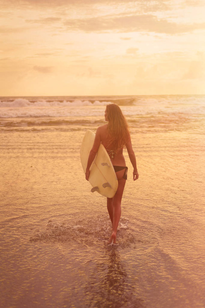 A young woman in bikini stands with her back to the camera and holds a surfboard by the ocean. Warm tinting - 写真・画像