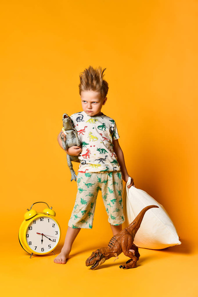 Little boy of preschool age with a pillow in his hands and a toy dinosaur waking up on an alarm clock. Photo in studio on a yellow background - Photo, Image