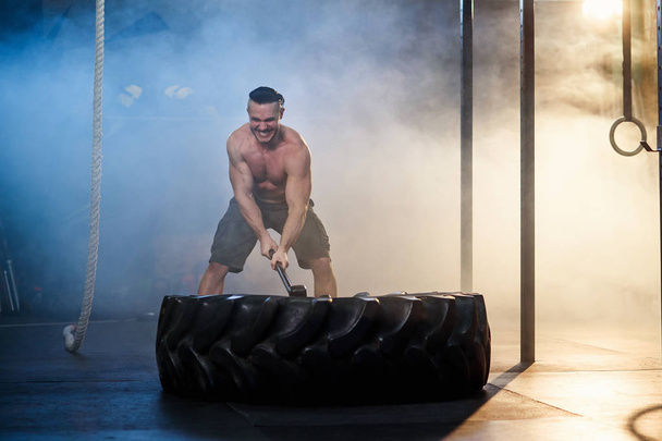 Attractive sport man hitting wheel tire with hammer during training. - Photo, Image