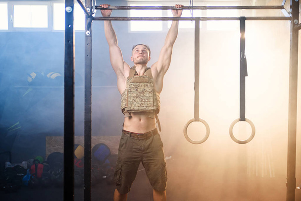 Young sports man with heavy waistcoat makes pull ups on crossbar in the gym full of smoke. - Foto, Imagem