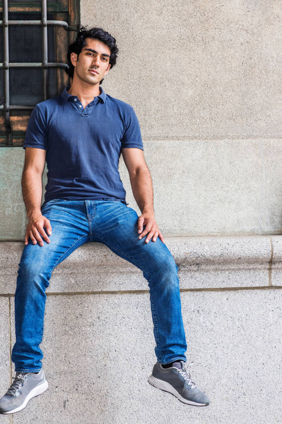 Портрет студента американского колледжа Young Mix Race в Нью-Йорке, одетого в синюю рубашку с короткими рукавами, синие джинсы, сидящего у стены старого стиля на улице в кампусе, расслабляющегося, мыслящего
 - Фото, изображение