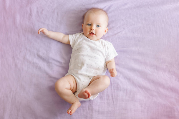 Πορτραίτο του χαριτωμένο αξιολάτρευτο χαμόγελο λευκό καυκάσιο μωρό κορίτσι με μπλε μάτια τέσσερις μήνες παλιά ξαπλωμένοι στο κρεβάτι κοιτάζοντας την κάμερα. Δείτε από πάνω. Ευτυχισμένος παιδικός τρόπος ζωής. - Φωτογραφία, εικόνα
