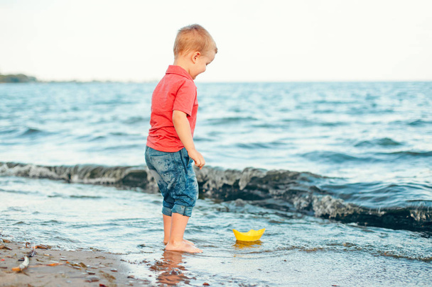 Happy Caucasian red-haired toddler child boy putting yellow paper boat in water on lake sea ocean shore at evening sunset or morning. Kid playing on beach. Happy lifestyle childhood concept. - Photo, image