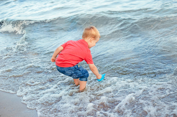 Happy Caucasian red-haired toddler child boy putting blue paper boat in water on lake sea ocean shore at evening sunset or morning. Kid playing on beach. Happy lifestyle childhood concept. - Fotó, kép