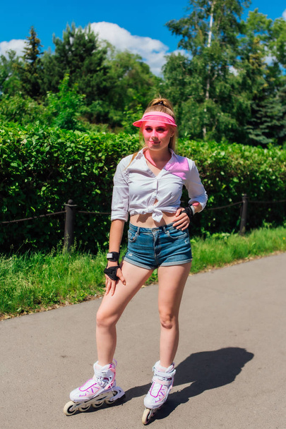 Portrait of an emotional girl in a pink cap visor and protective gloves for rollerblades and skateboarding riding on rollerblades on the road. - Zdjęcie, obraz