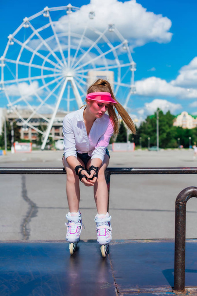 Portrait of an emotional girl in a pink cap visor wearing protective gloves and rollerblades sitting on the background of ferris wheel. - Photo, Image