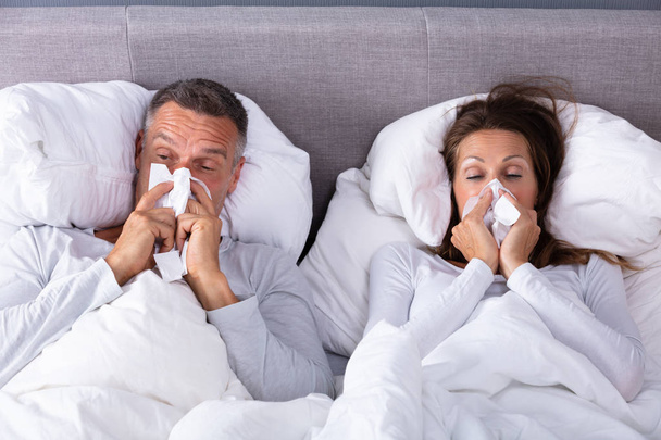 Close-up Of Mature Couple Lying On Bed Blowing Their Nose In Bedroom - Valokuva, kuva