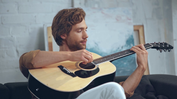 молодой улыбающийся мужчина играет на акустической гитаре на диване
 - Кадры, видео