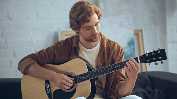 koncentrált fiatalember, akusztikus gitárt játszani a kanapén - Felvétel, videó