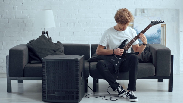 スピーカーの近くのソファでエレキギターを演奏する若いロッカー - 映像、動画