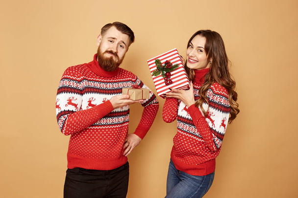 Gelukkig meisje en een man gekleed in rode en witte truien met herten houden Kerstcadeaus in hun handen op een beige achtergrond in de Studio - Foto, afbeelding