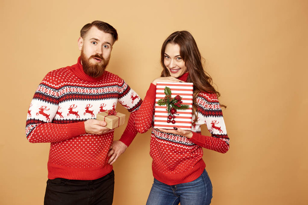 Happy girl and a guy dressed in red and white sweaters with deer hold Christmas gifts in their hands on a beige background in the studio - Photo, image