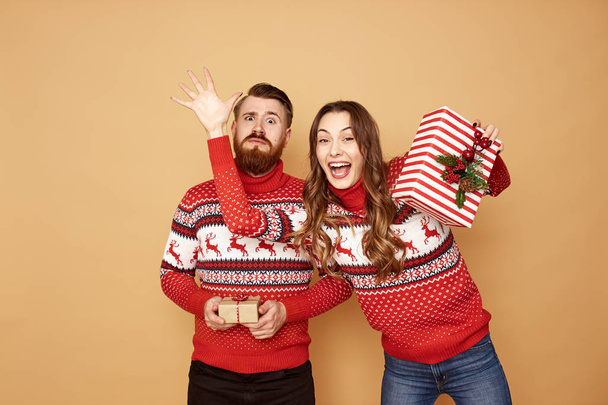 Happy girl and a guy dressed in red and white sweaters with deer hold Christmas gifts in their hands on a beige background in the studio - Fotografie, Obrázek