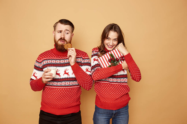Girl holding Christmas gift and a guy holding a cookie and a glass of milk dressed in red and white sweaters with deer stand on a beige background in the studio - Foto, Imagen