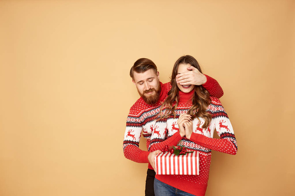 Guy closes the girls eyes with his hand and givs her a gift on a beige background in the studio.They both dressed in red and white sweaters with deer. - Zdjęcie, obraz